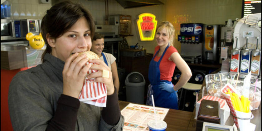 Zicht op toog met twee dames in blauwe schort en één dame op de voorgrond die een hap neemt van een hamburger.