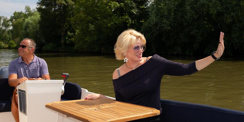 Pascale Platel met zonnebril die in een boot zit en naar iemand zwaait, de bootbestuurder