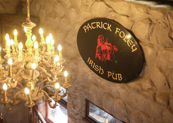 Reclamebord op zwarte achtergrond, met de letters 'Patrick Foley' en 'Irish Pub' in goud. In het midden staat een vioolspeler in het rood afgebeeld.