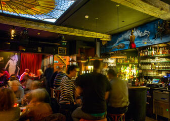 binnenkant bar, veel flessen tegen de muur, mensen aan tafeltjes, schilderij zingende vrouw, boot en gitaar op de zee