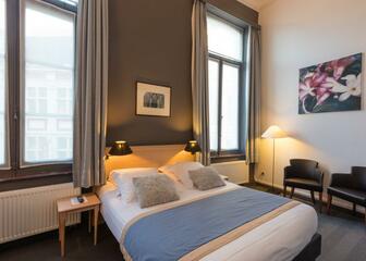 hotelkamer met tweepersoonsbed, 2 zwarte stoelen, grote ramen met grijze gordijnen