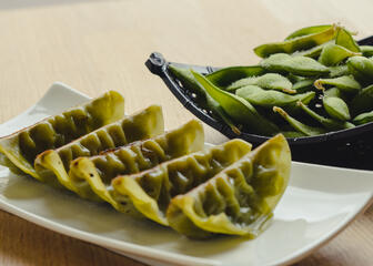 5 grüne Gyōza auf einem weißen Teller mit Edamame in einer schwarzen Schale