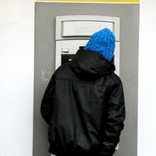 Man wearing black jacket, blue cap and pair of jeans is taking wallet out of his back pocket. He's standing in front of an ATM with a yellow sign with blue letters saying 'CASH'.